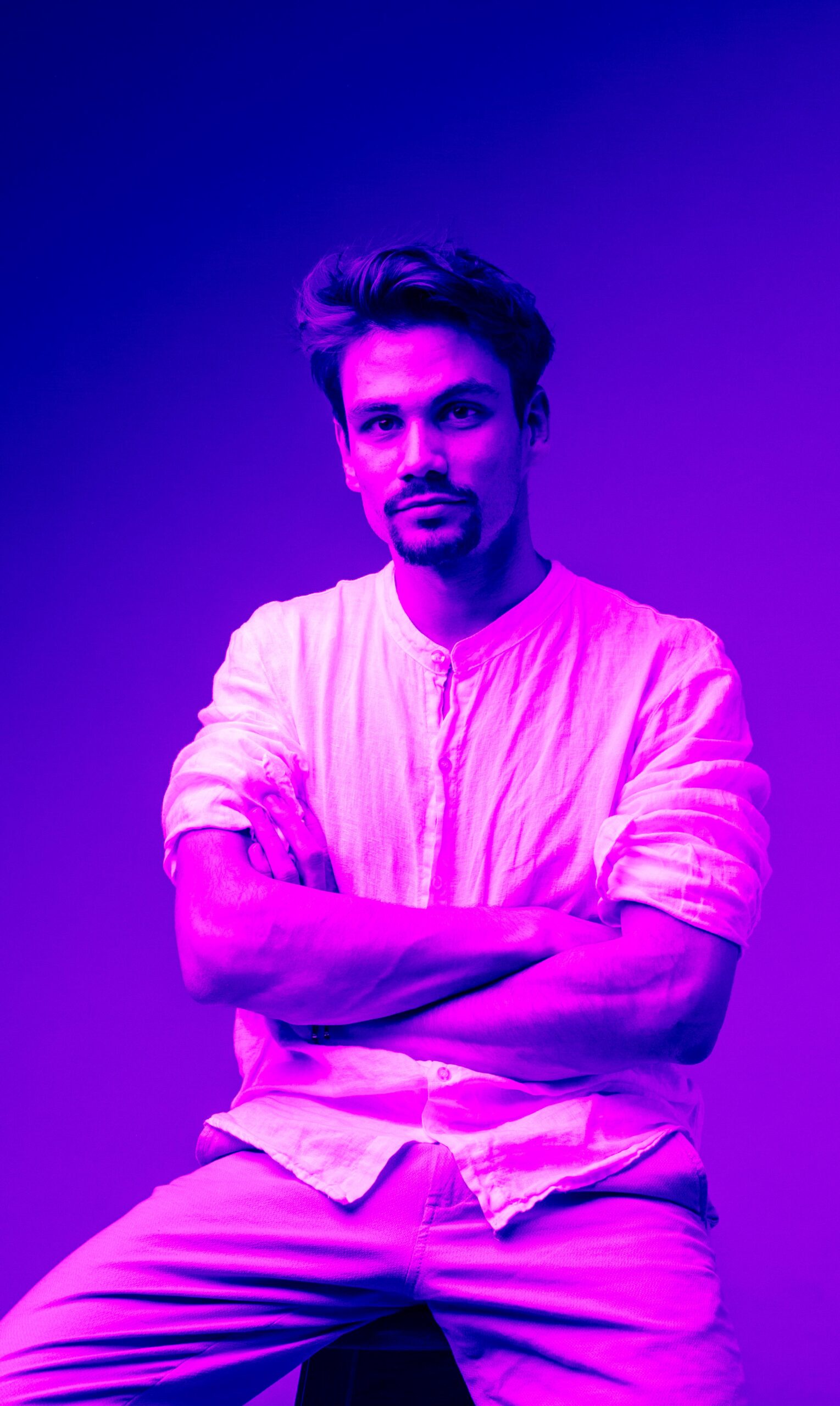 Malumo Portret Malumo visual artist portrait in a white shirt, intensely lit with vibrant purple and deep blue neon studio lighting.